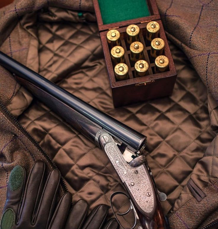 Shotgun with engraved metalwork rests on a brown quilted coat, beside a wooden box of brass shotgun shells and hunting gloves.