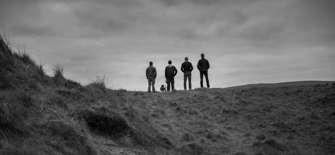 Four people and a dog stand on a grassy hill, looking toward a cloudy horizon.